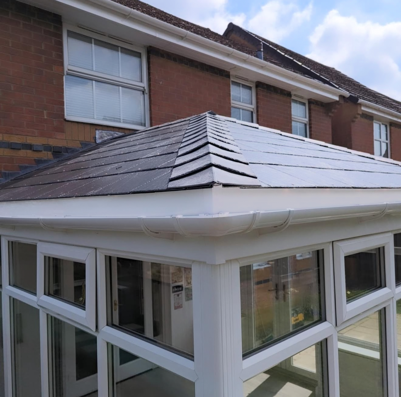 Close-up of a conservatory with a dark slate roof, central ridge and white guttering against a brick house