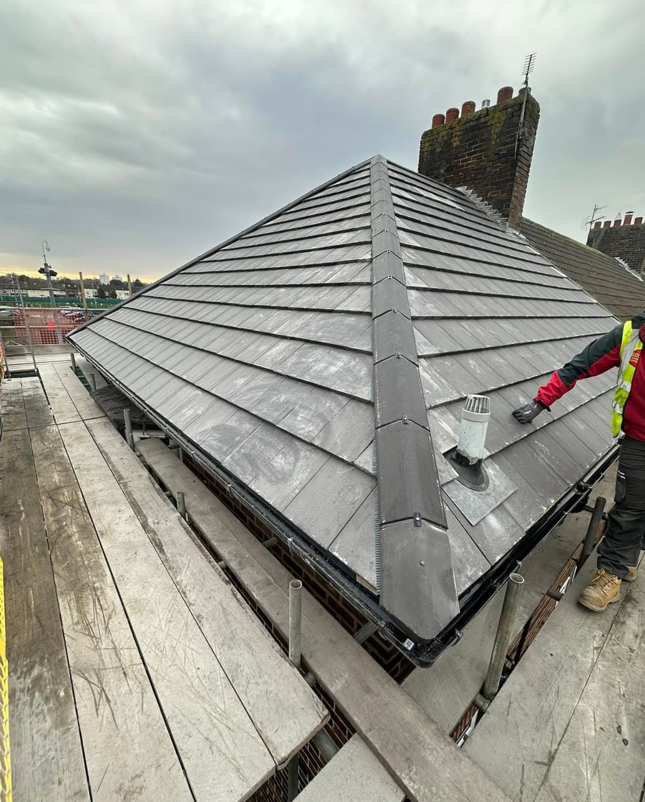 Grey tiled roof with central ridge, multiple brick chimneys and a roofer finishing the detailing
